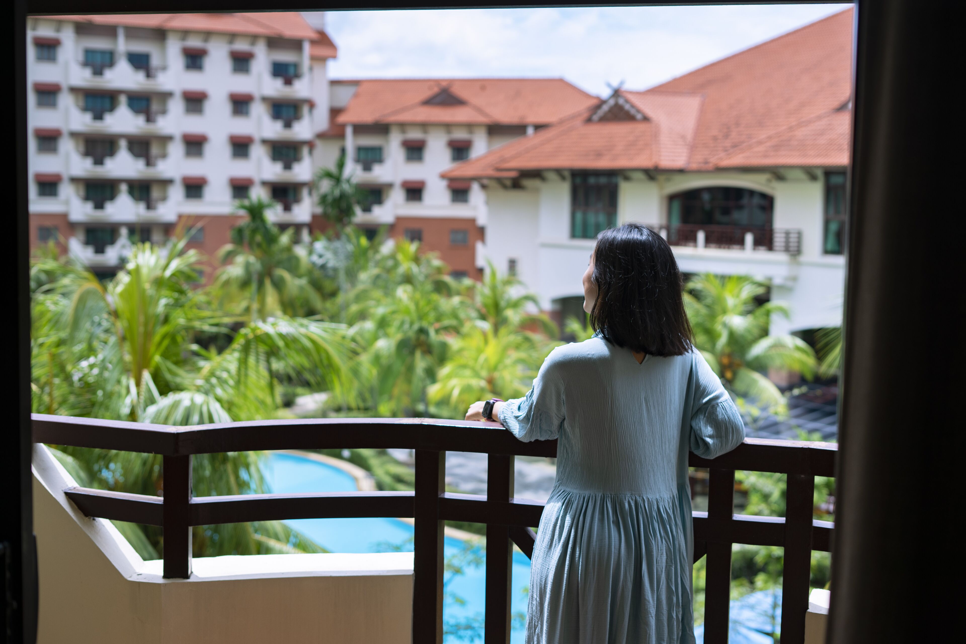 A person standing on a balcony overlooking a pool and lush greenery, with a building featuring balconies and a red roof in the background.