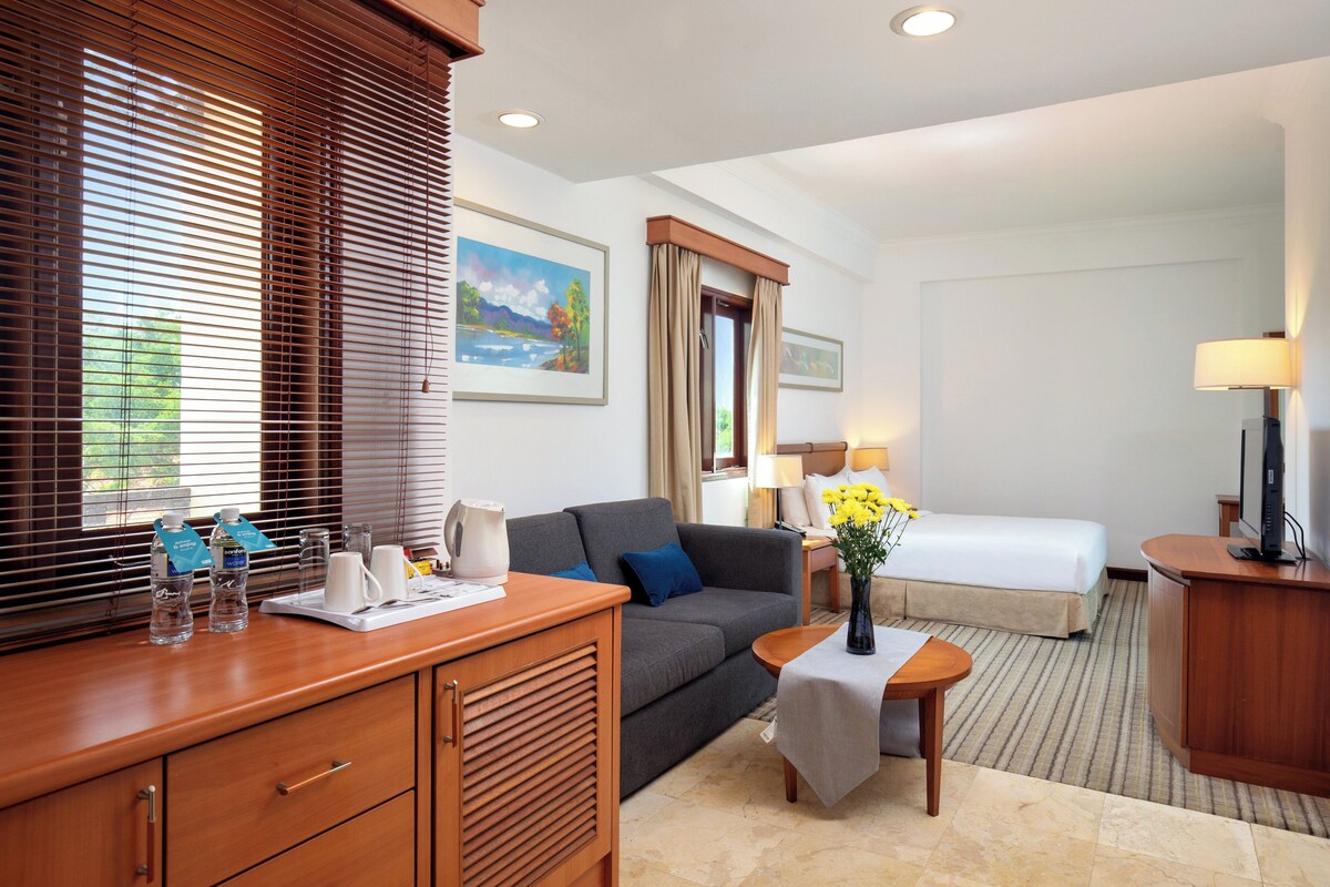 A hotel room with a sofa, a small table with a vase of flowers, a bedside table with lamps, a bed, and a desk with a computer.