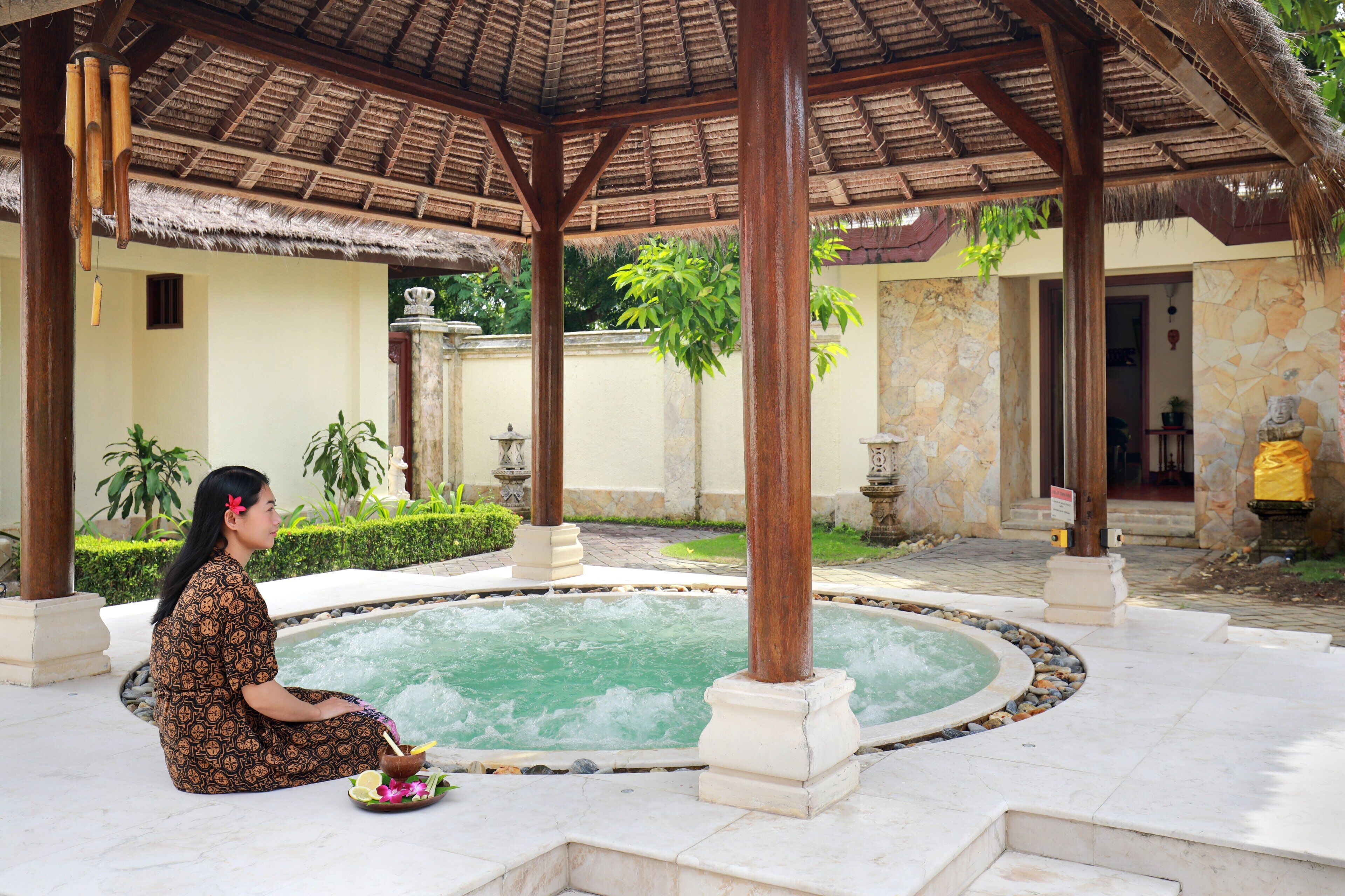 A woman sitting by a circular water feature in a covered outdoor area with stone walls and a thatched roof.