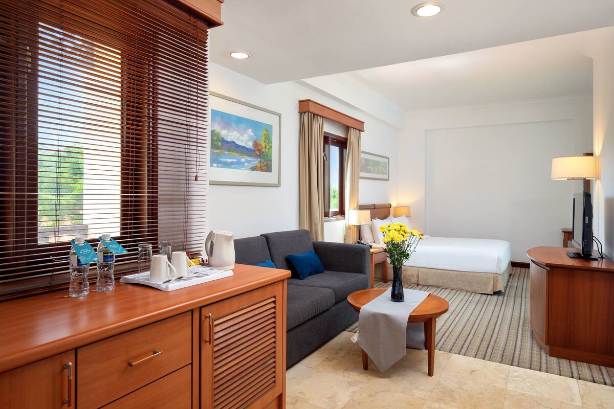 Habitación de hotel con sofá, una mesita con un florero, mesitas de noche con lámparas, cama y escritorio con computadora.