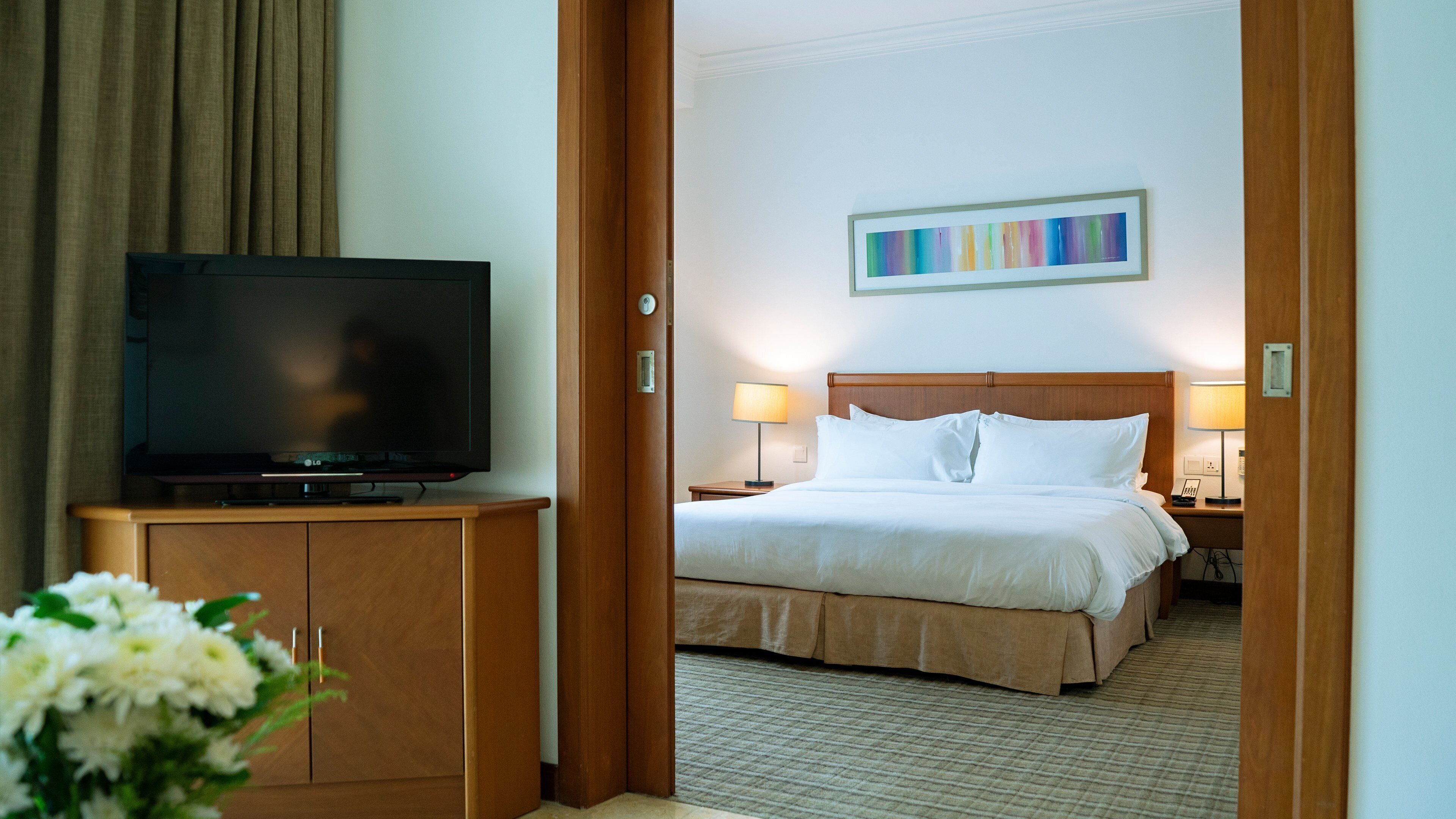 Une chambre d’hôtel avec un lit, un téléviseur posé sur une commode en bois, un tableau au mur et un arrangement floral.