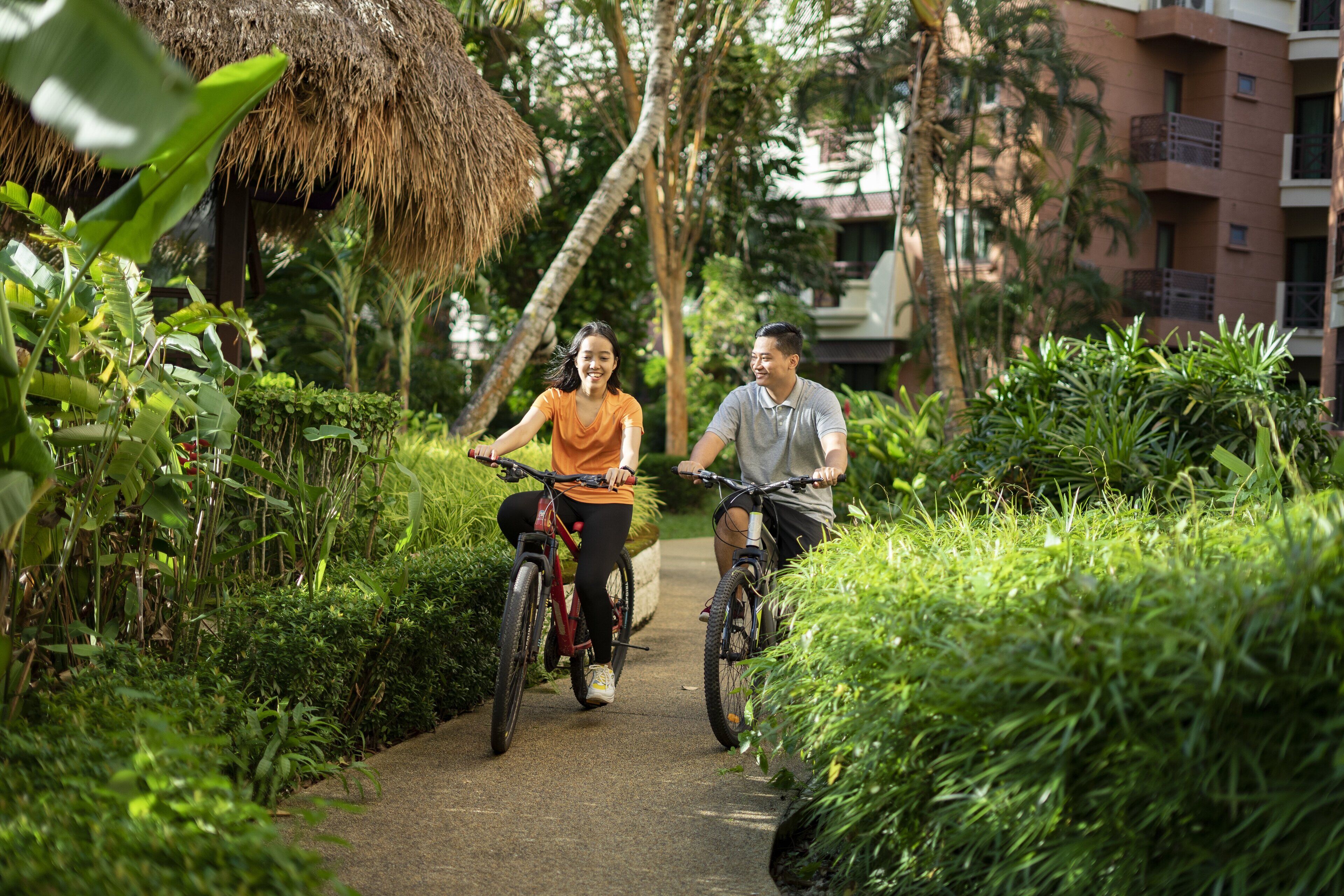 Two people riding bicycles on a pathway surrounded by lush greenery and a thatched-roof structure.