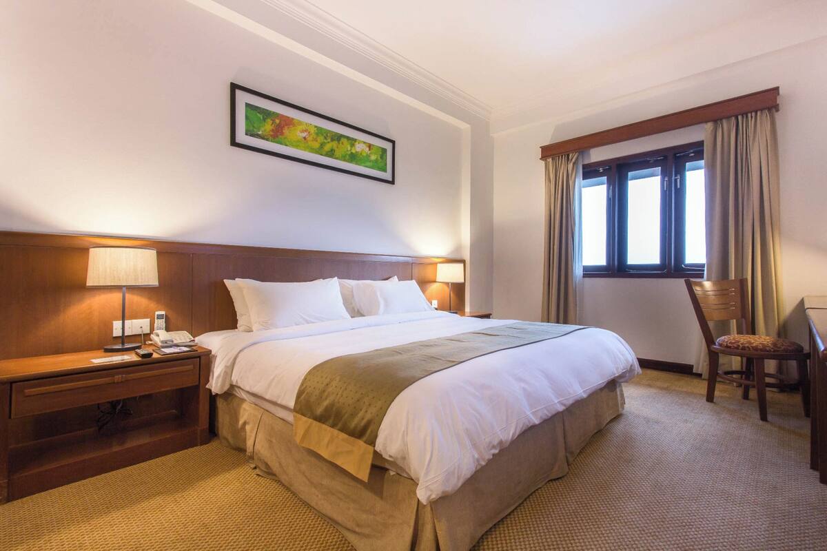 Una habitación de hotel con una cama grande, dos mesitas de noche con lámparas, una silla, una ventana con cortinas y un cuadro en la pared.