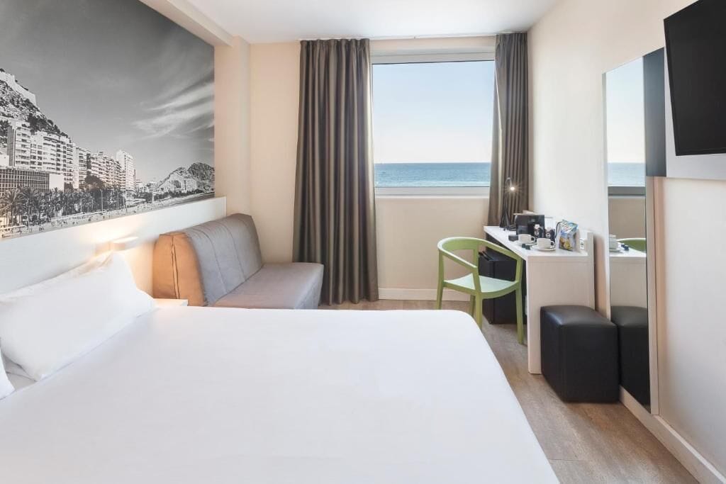 Habitación de hotel con cama, escritorio, silla, sofá, un gran ventanal con cortinas y un mural de paisaje urbano en la pared.