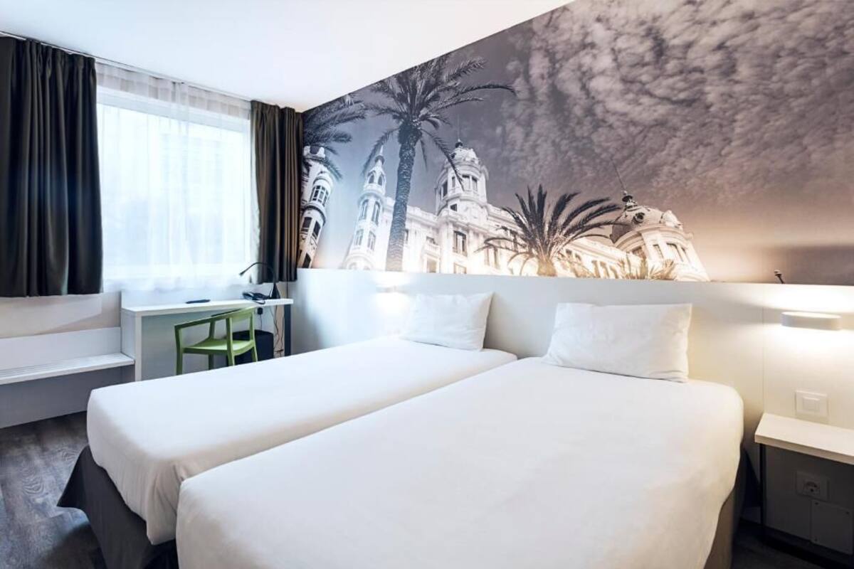 A hotel room with two beds, a desk, a chair, a lamp, and a mural of a building with palm trees.