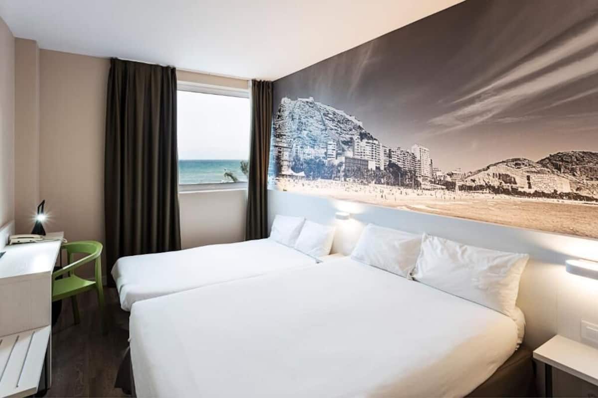 A hotel room with a bed, a desk, a chair, and a large window with a beach view.