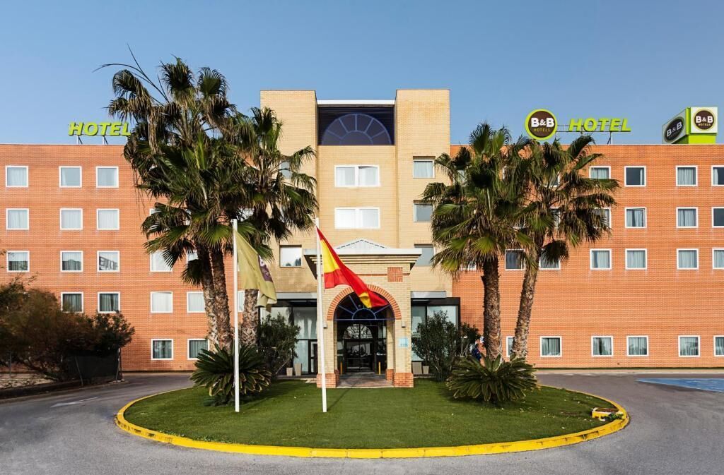 Un hôtel à la façade en briques, doté de nombreuses fenêtres et bordé de palmiers au premier plan.