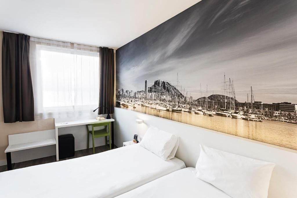 A hotel room with a bed, a desk, a chair, and a large cityscape painting on the wall.
