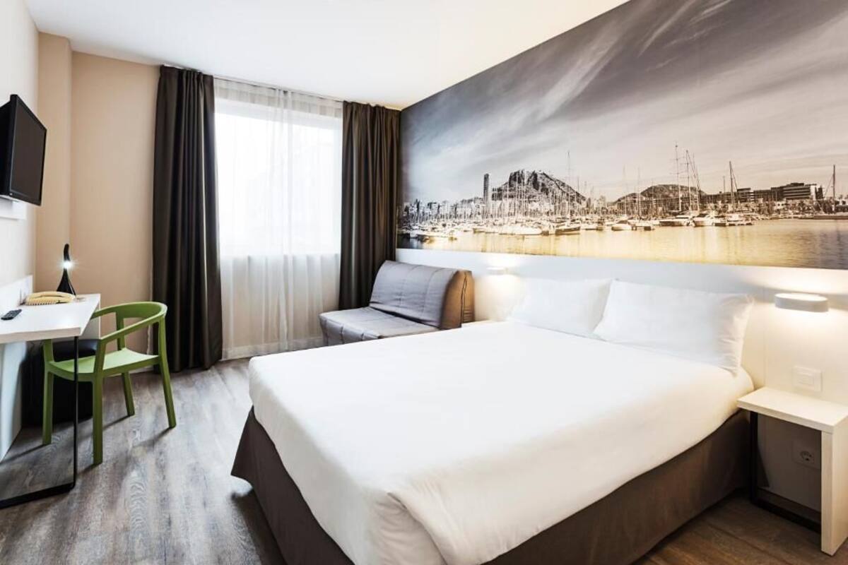 A hotel room with a bed, a desk, a chair, a TV, and a cityscape mural on the wall.