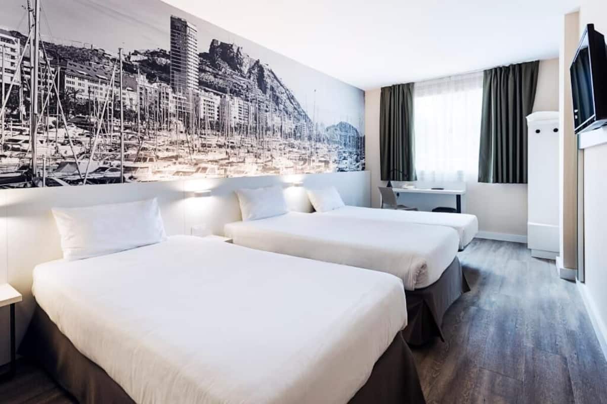 A hotel room with two beds, a desk, a TV, and a large cityscape mural on the wall.