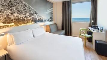 A hotel room with a bed, a desk, a chair, and a large window with a view of the sea.