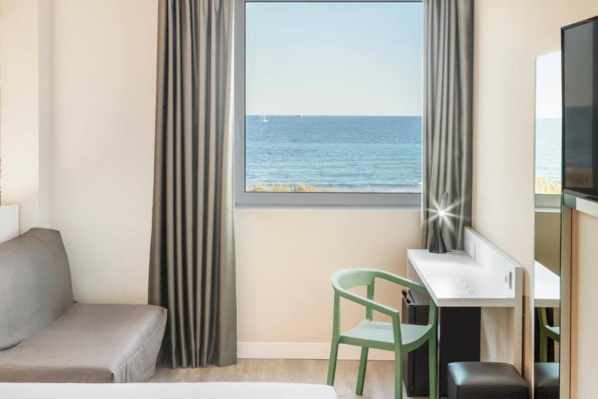 A hotel room with a bed, a desk, a chair, a TV, and a view of the sea.