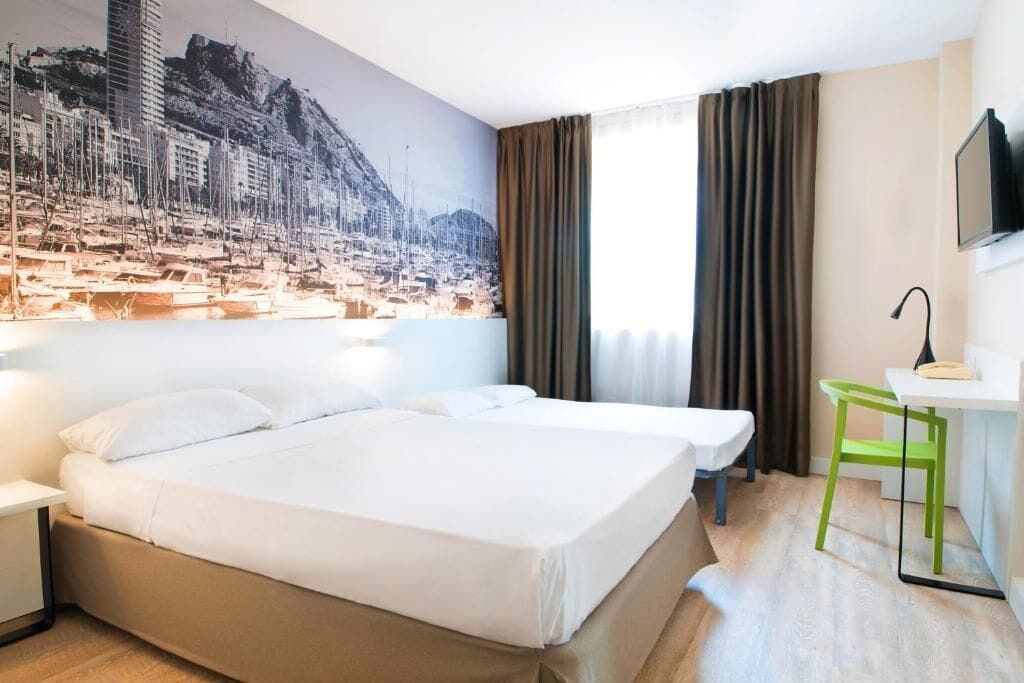 A hotel room with a bed, a desk, a chair, a television, and a large cityscape mural on the wall.