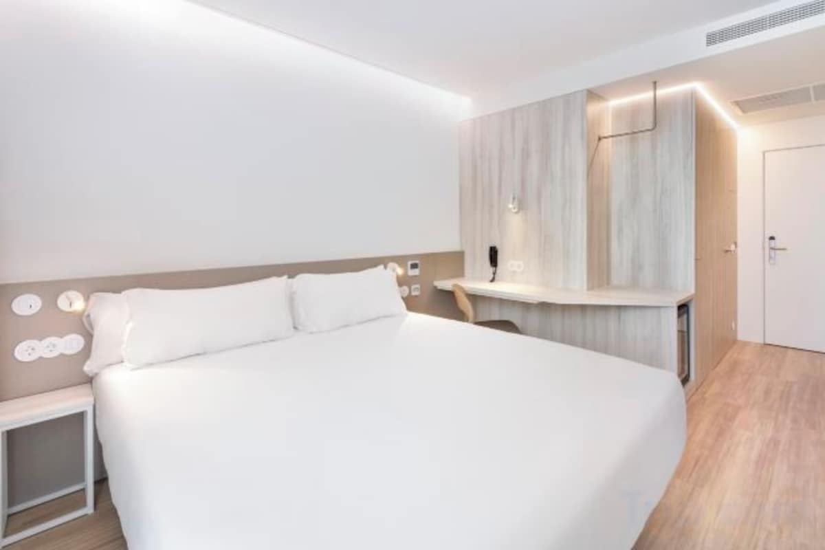 A modern hotel room with a large bed, bedside table, and a built-in desk with a chair.