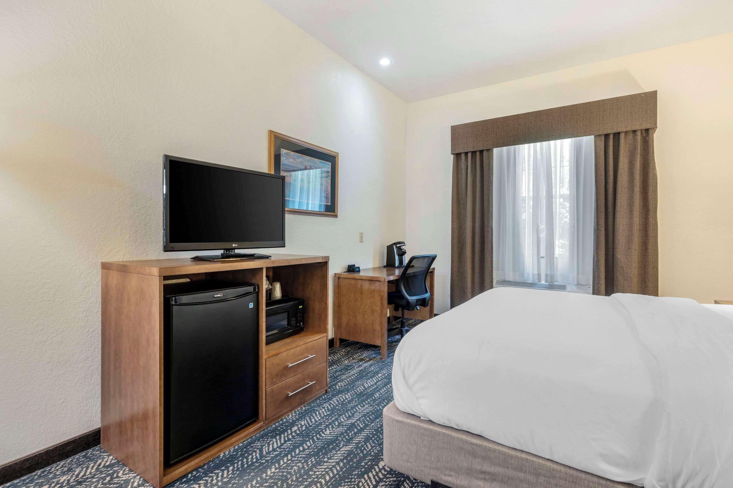 A hotel room with a bed, a desk with a chair, a television, a refrigerator, and a window with curtains.