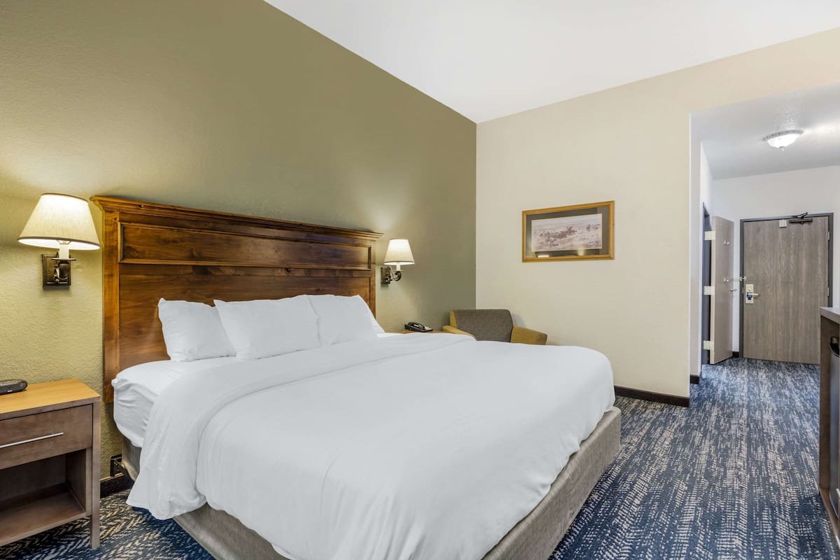 A hotel room with a large bed, two bedside lamps, a chair, a desk, and a door leading to another room.