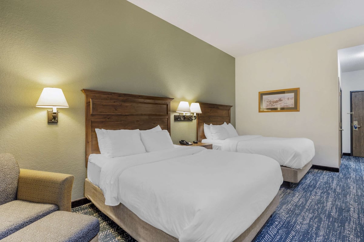 A hotel room with two beds, a chair, a nightstand, and a wall-mounted lamp.