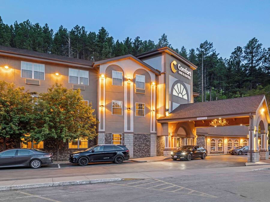 Un hotel Comfort Inn & Suites con entrada cubierta, varios coches estacionados al frente y árboles en el fondo.