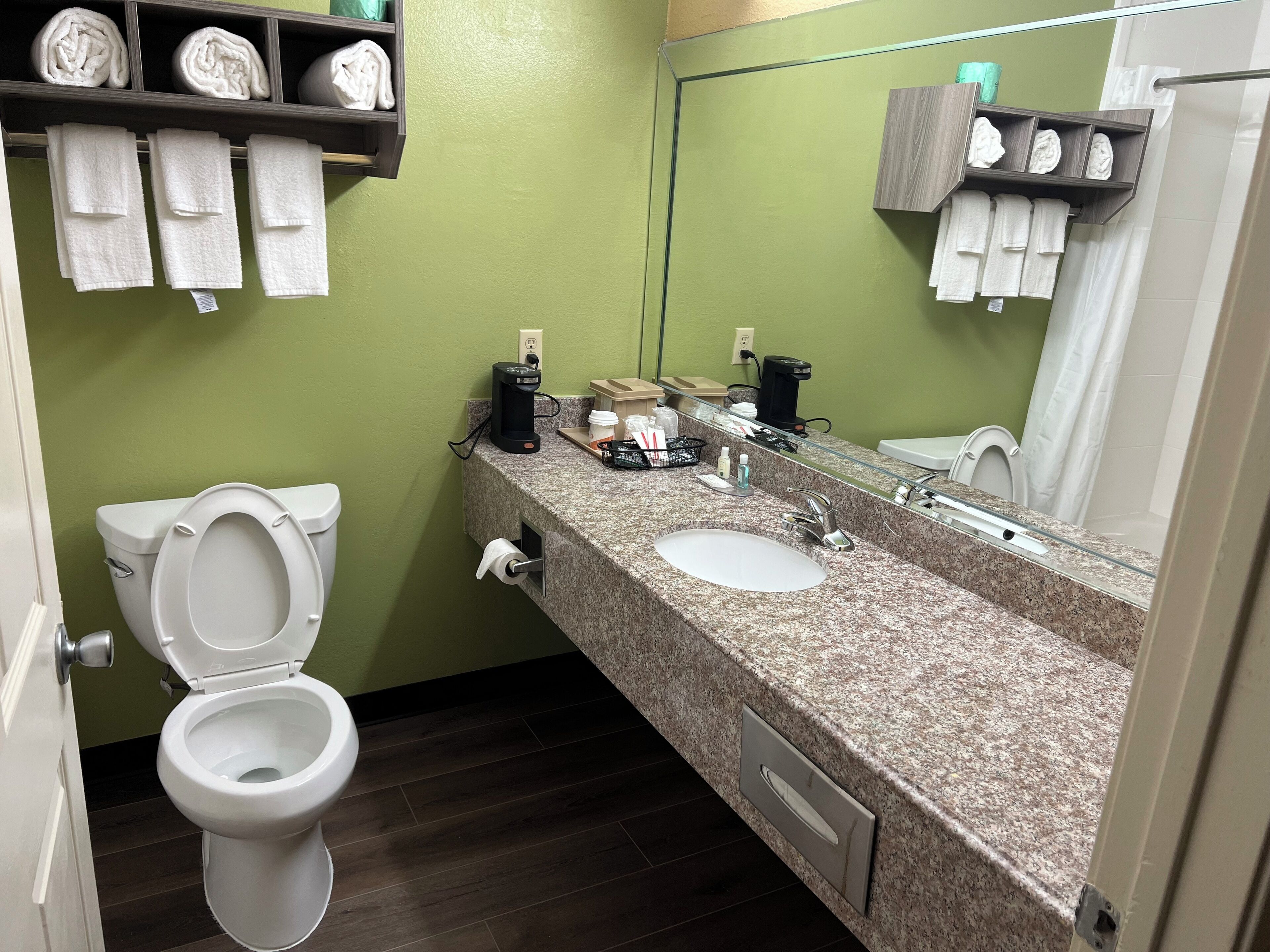 A bathroom with a toilet, sink, and a mirror. There are towels on a shelf and a coffee maker on the counter.