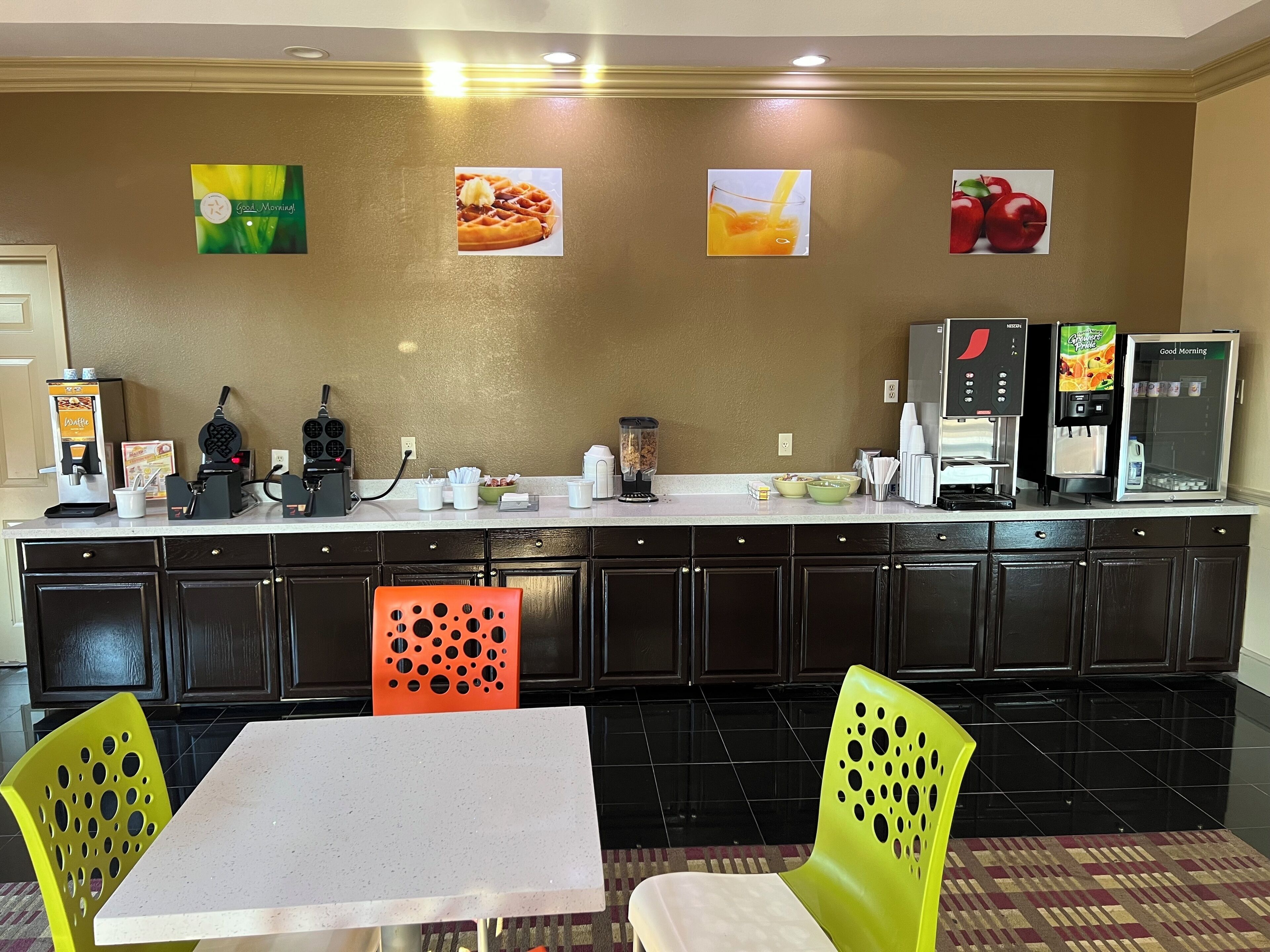 A hotel kitchen with a buffet setup, including a waffle maker, coffee machine, and juice dispenser.