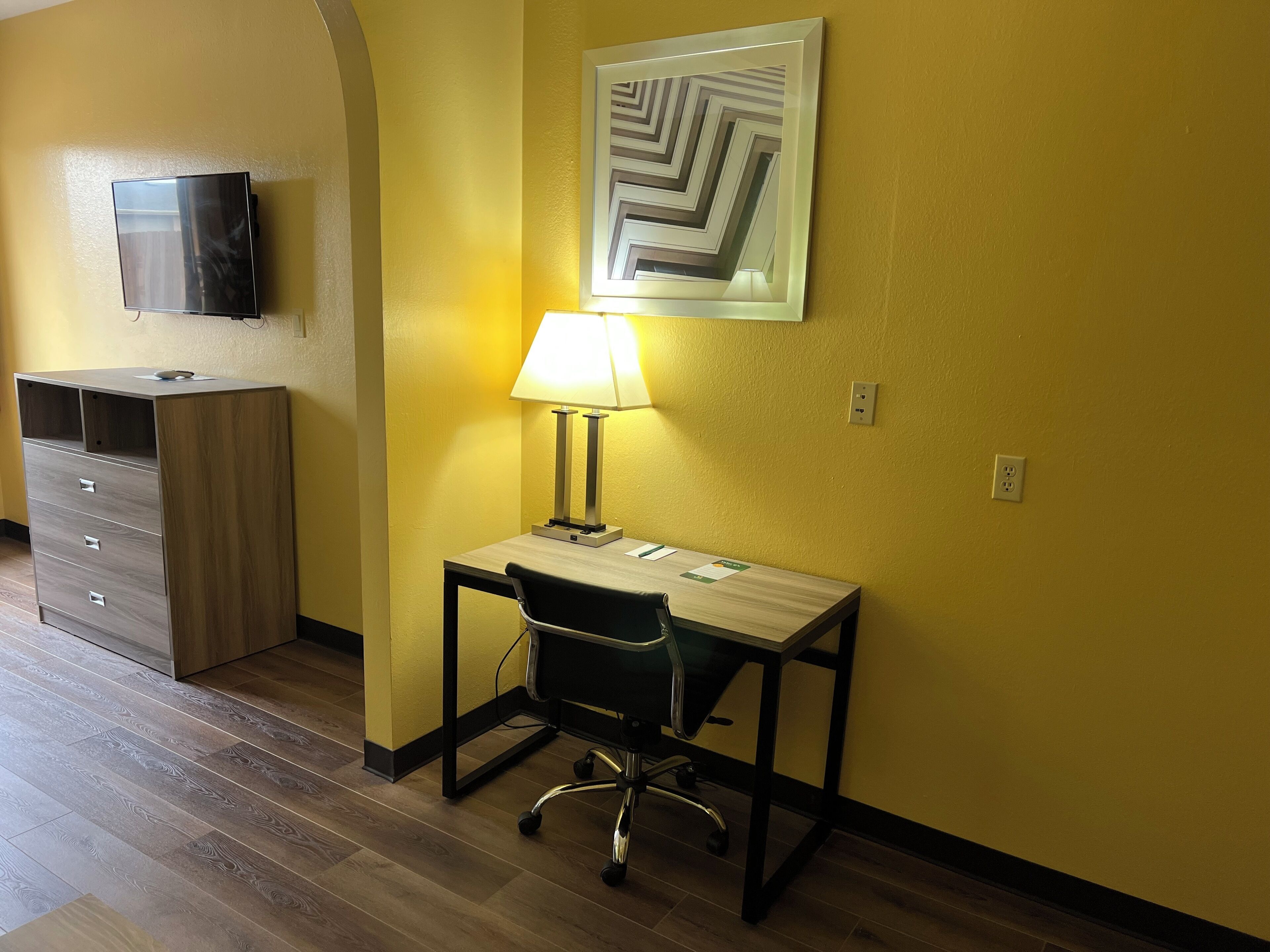 Una camera d'albergo con una scrivania, una sedia, una lampada e un televisore appeso al muro.
