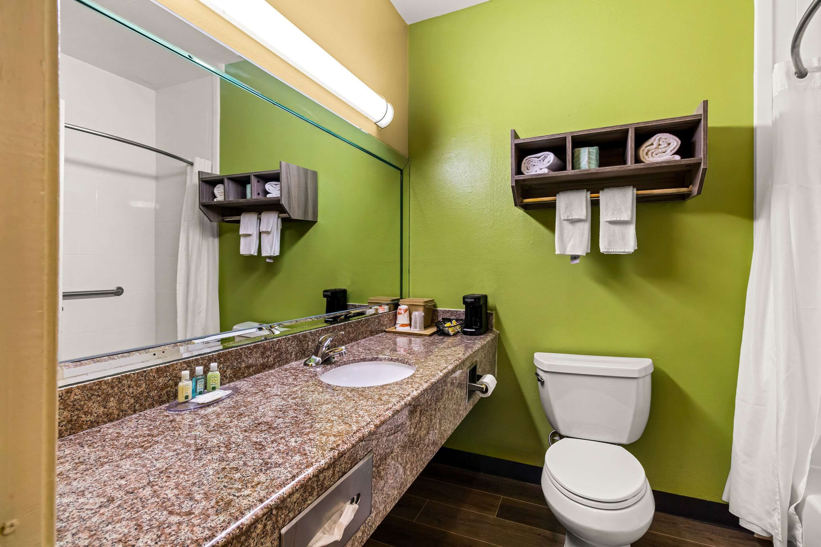 A bathroom with a granite countertop, a sink, a toilet, a mirror, and a shelf with towels.