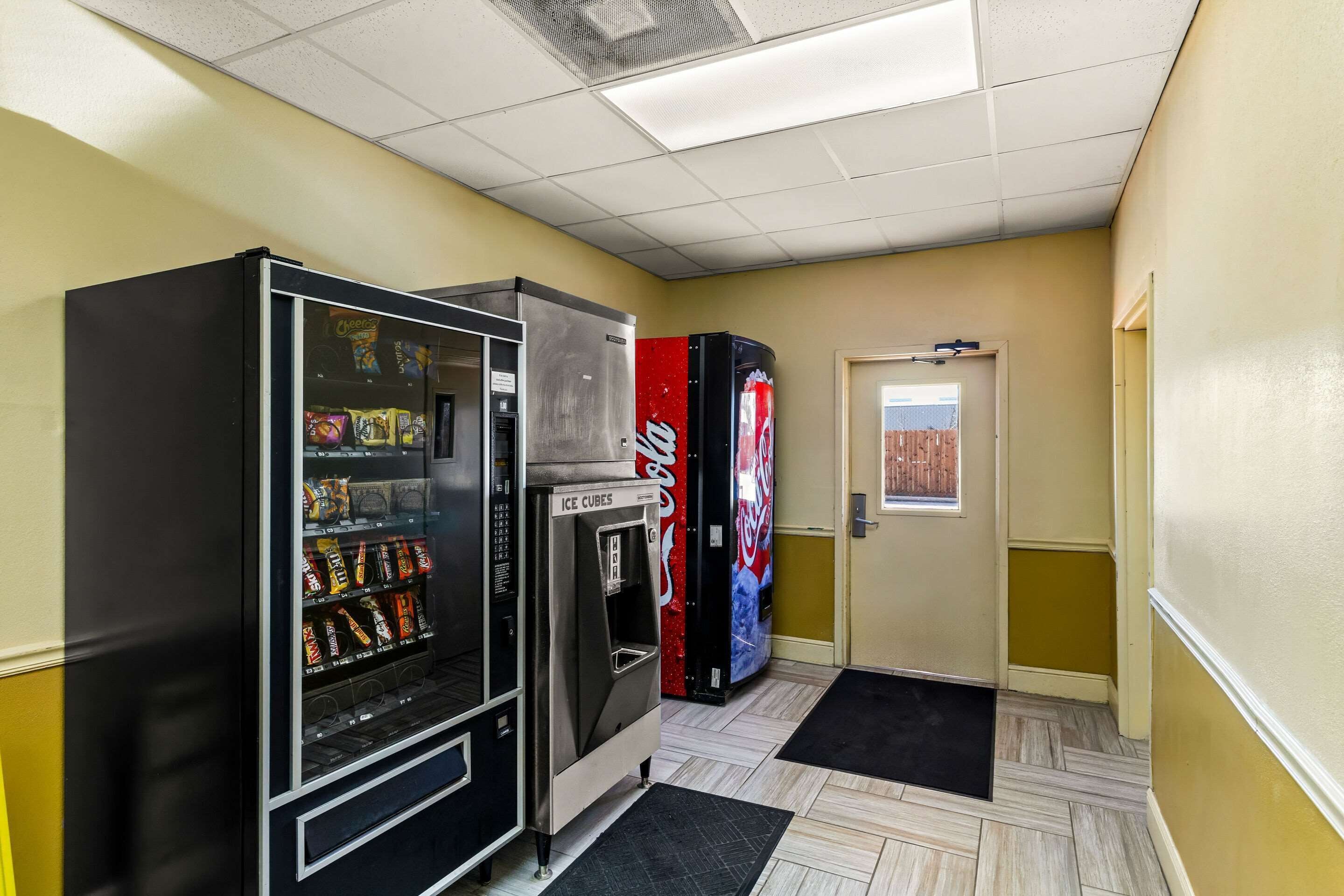 Un couloir avec des distributeurs automatiques, un réfrigérateur à boissons Coca-Cola et une porte donnant sur une vue d’une clôture.