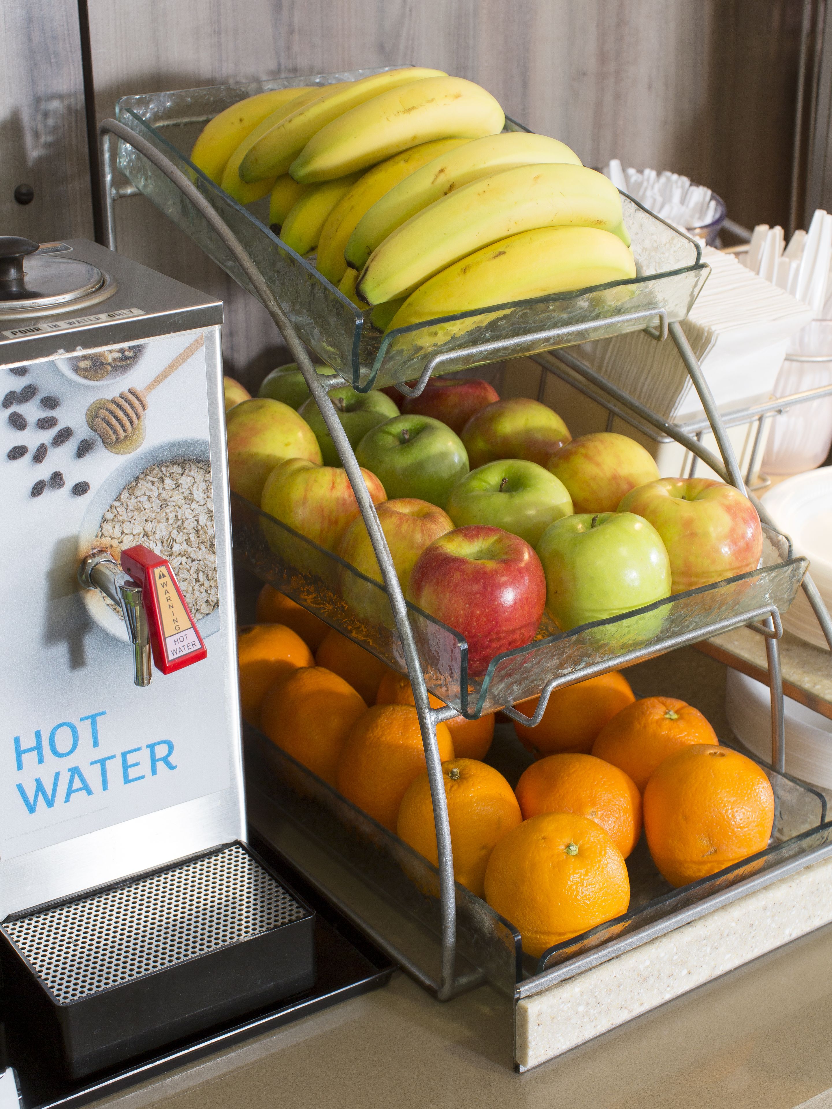 A self-service beverage station with a hot water dispenser, a fruit display with bananas, apples, and oranges, and a honey dipper.