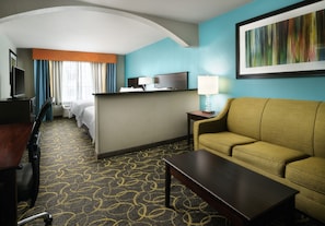 Quarto de hotel com cama, escrivaninha com cadeira, sofá, televisão e luminária.