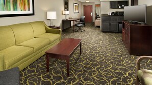 A hotel room with a green sofa, a wooden coffee table, a desk with a chair, a television, and a kitchen area with cabinets and a microwave. - Holiday Inn Express & Suites DFW - Grapevine by IHG (Grapevine)