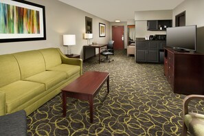 A hotel room with a green sofa, a wooden coffee table, a desk with a chair, a television, and a kitchen area with cabinets and a microwave. - Holiday Inn Express & Suites DFW - Grapevine by IHG (Grapevine)