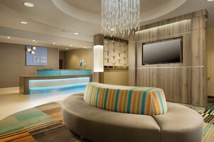 A hotel lobby with a reception desk, a television mounted on a wooden panel, a cushioned seating area, and a patterned carpet.