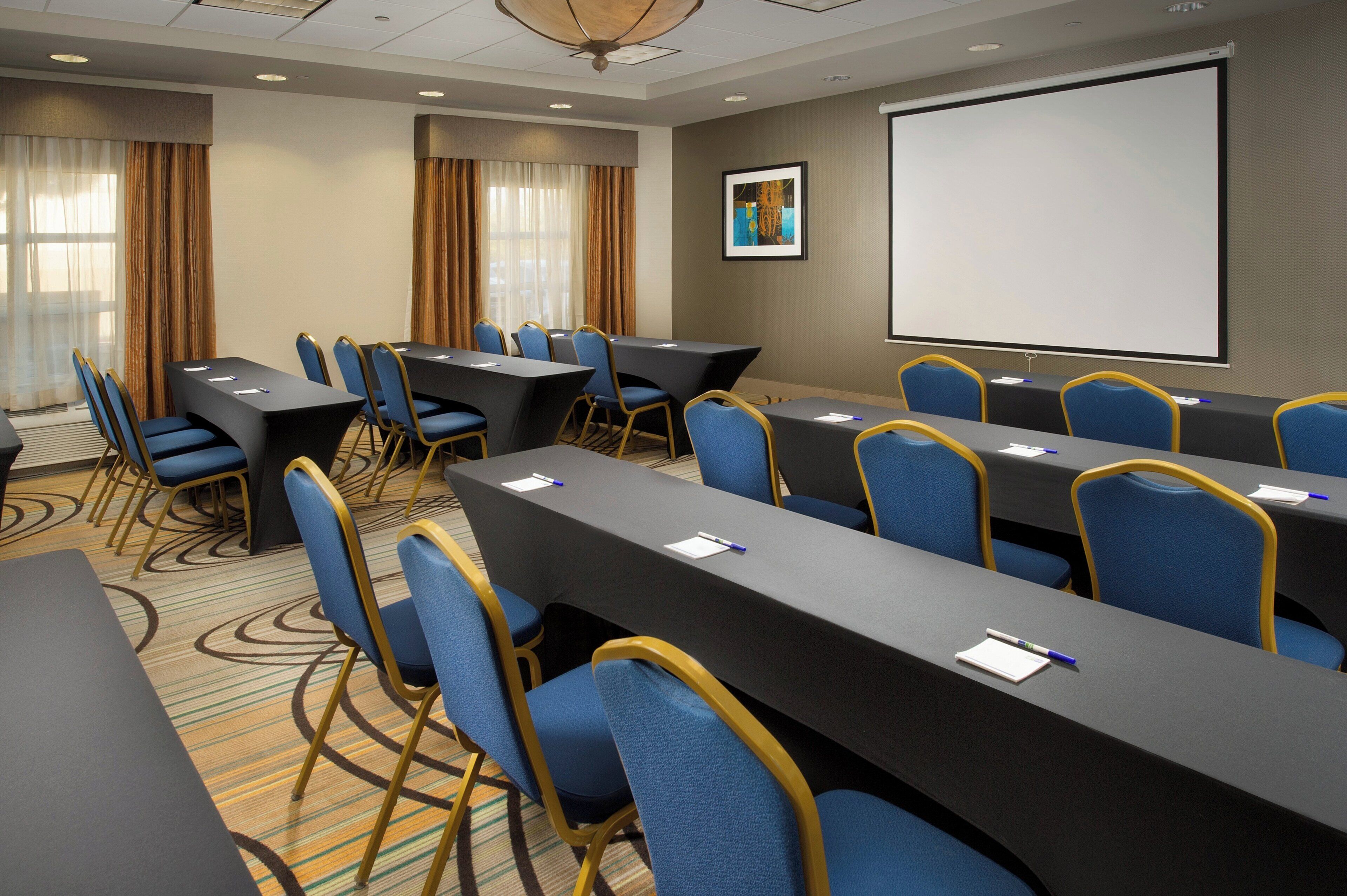 A conference room with long tables, blue chairs, a projector screen, and a painting on the wall.