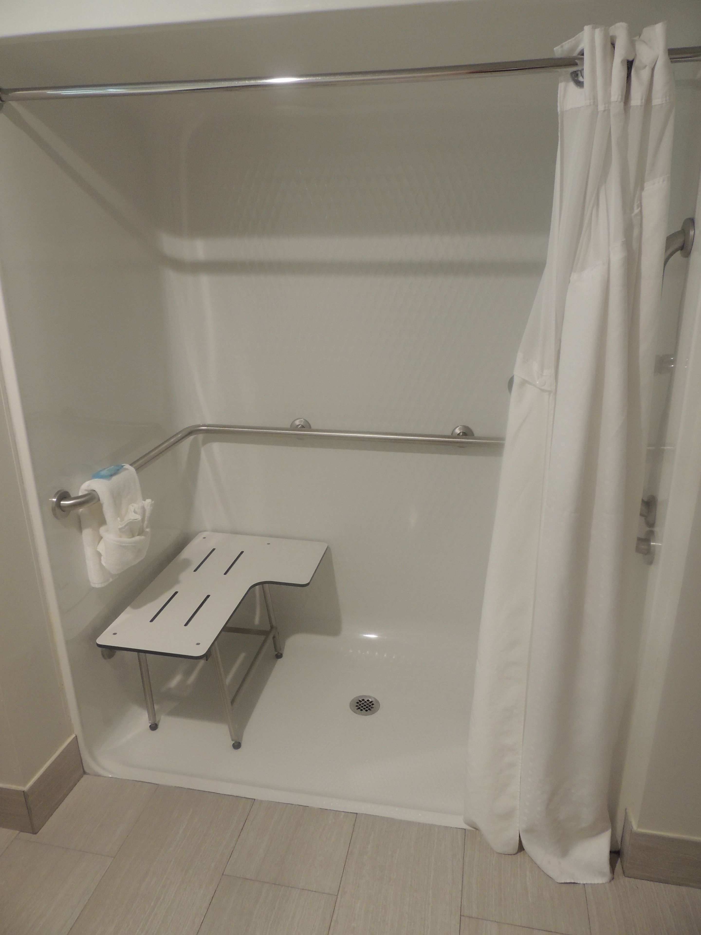 A shower area with a white shower curtain, a grab bar, a small white shelf, and a towel rack.