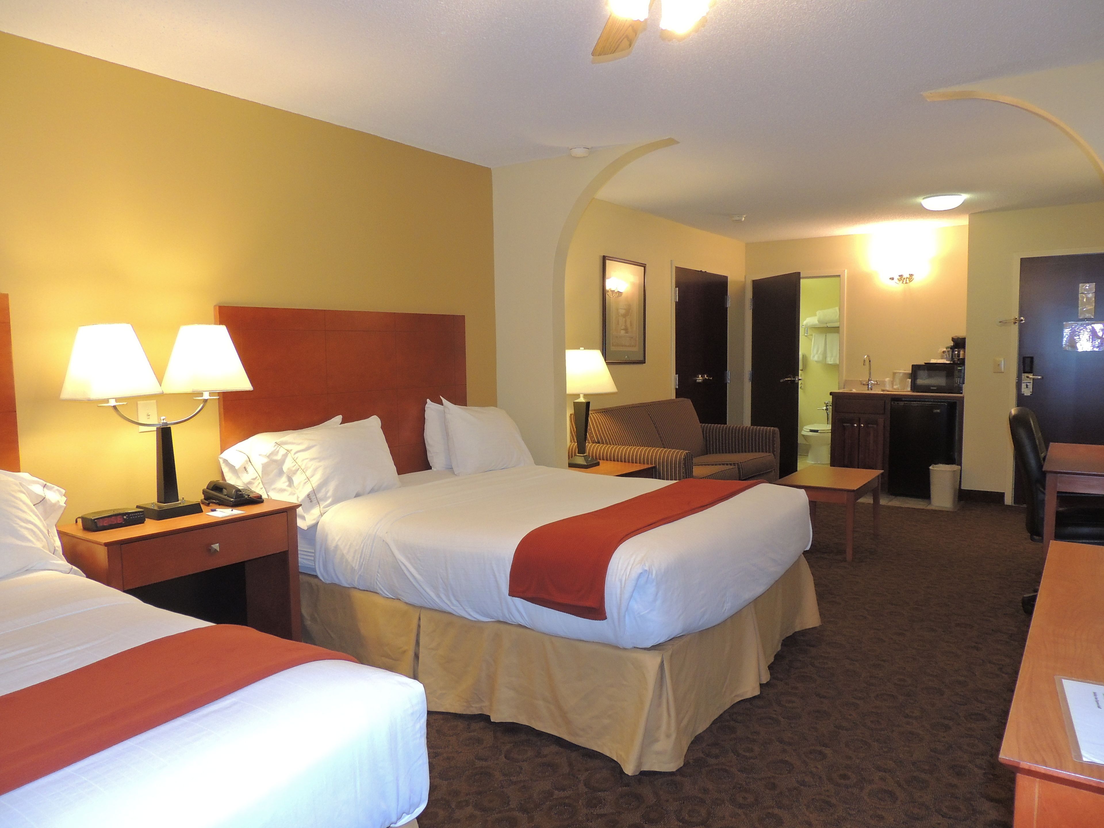A hotel room with two beds, a desk, a chair, a lamp, and a kitchenette.