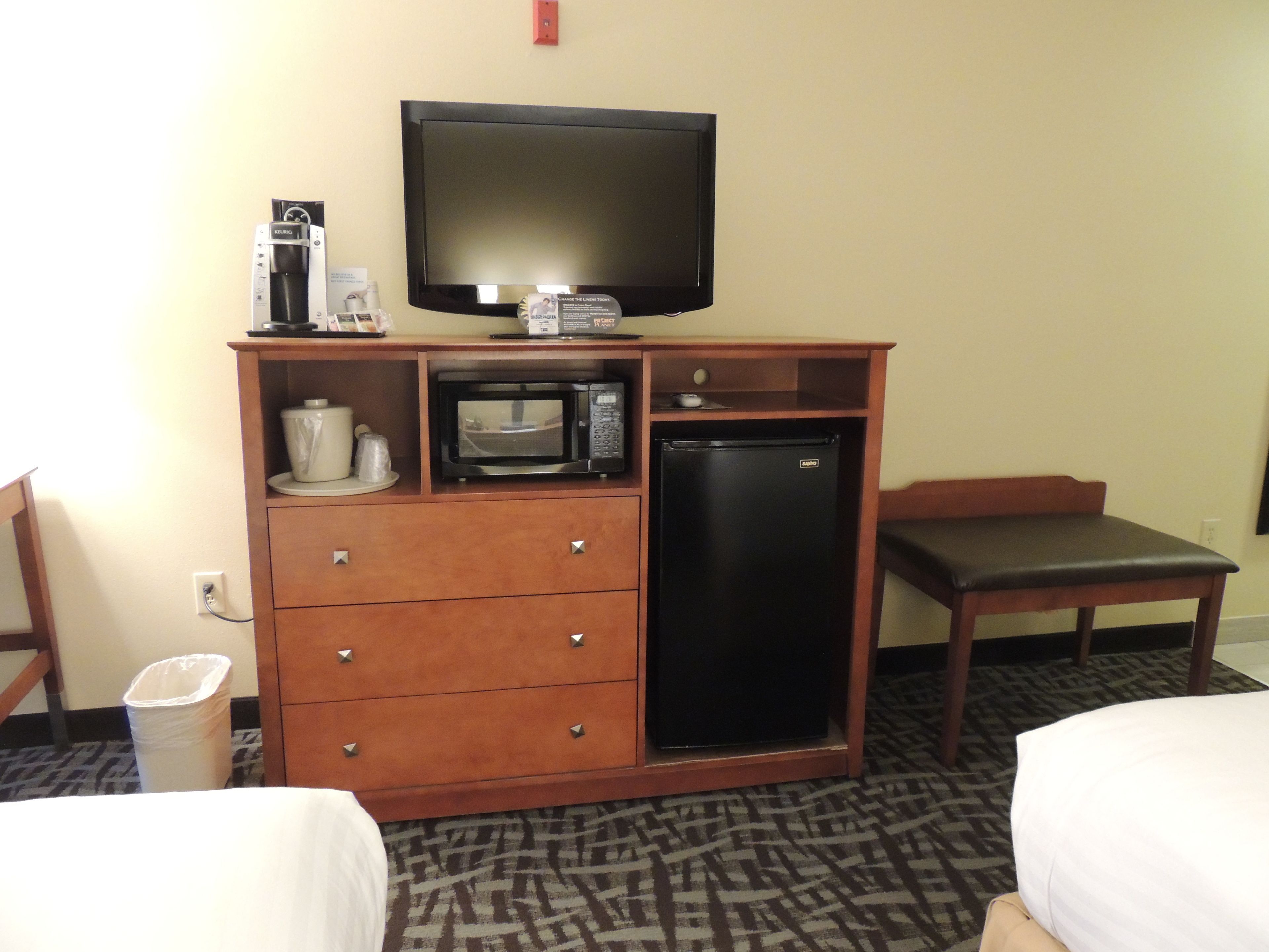 A hotel room with a wooden cabinet containing a microwave, a refrigerator, and a television. There is a chair and a bed in the room.