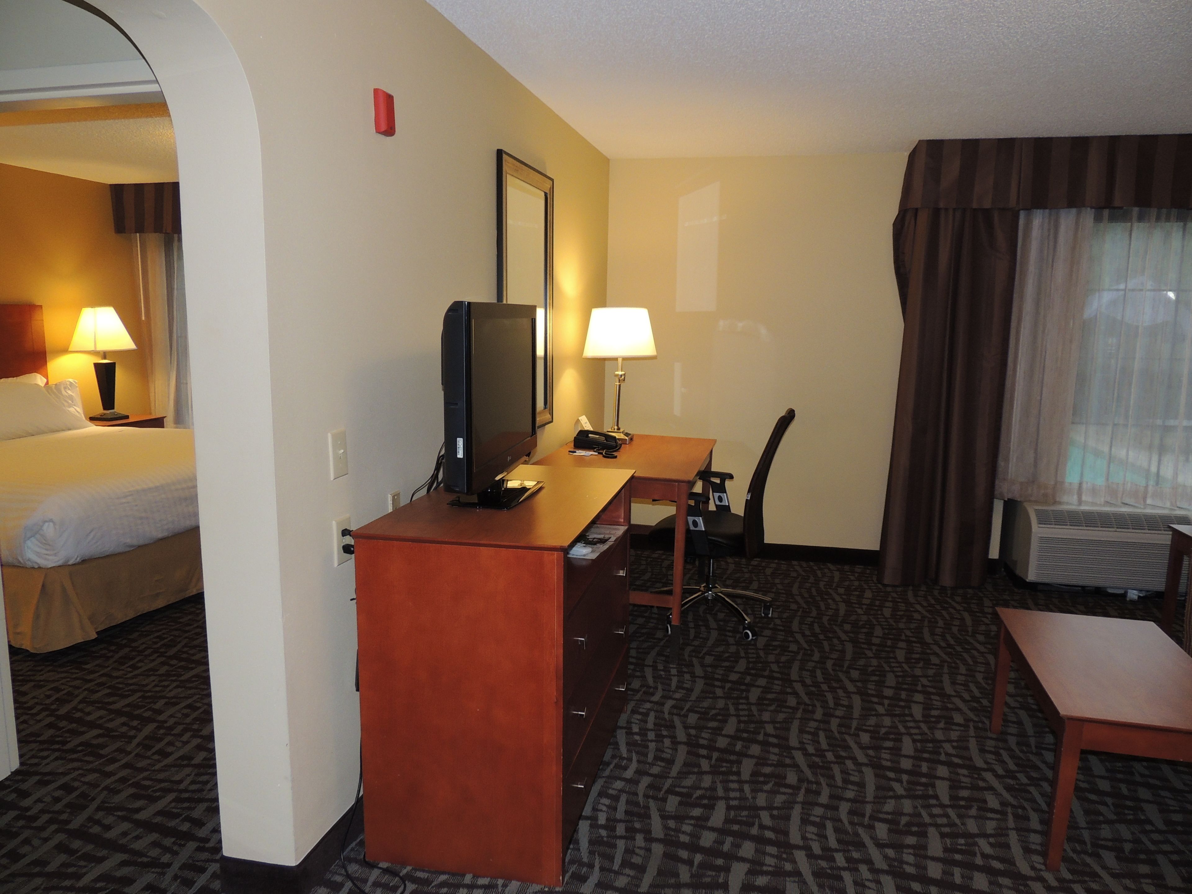 Quarto de hotel com cama, escrivaninha, cadeira, televisão e uma pequena mesa.