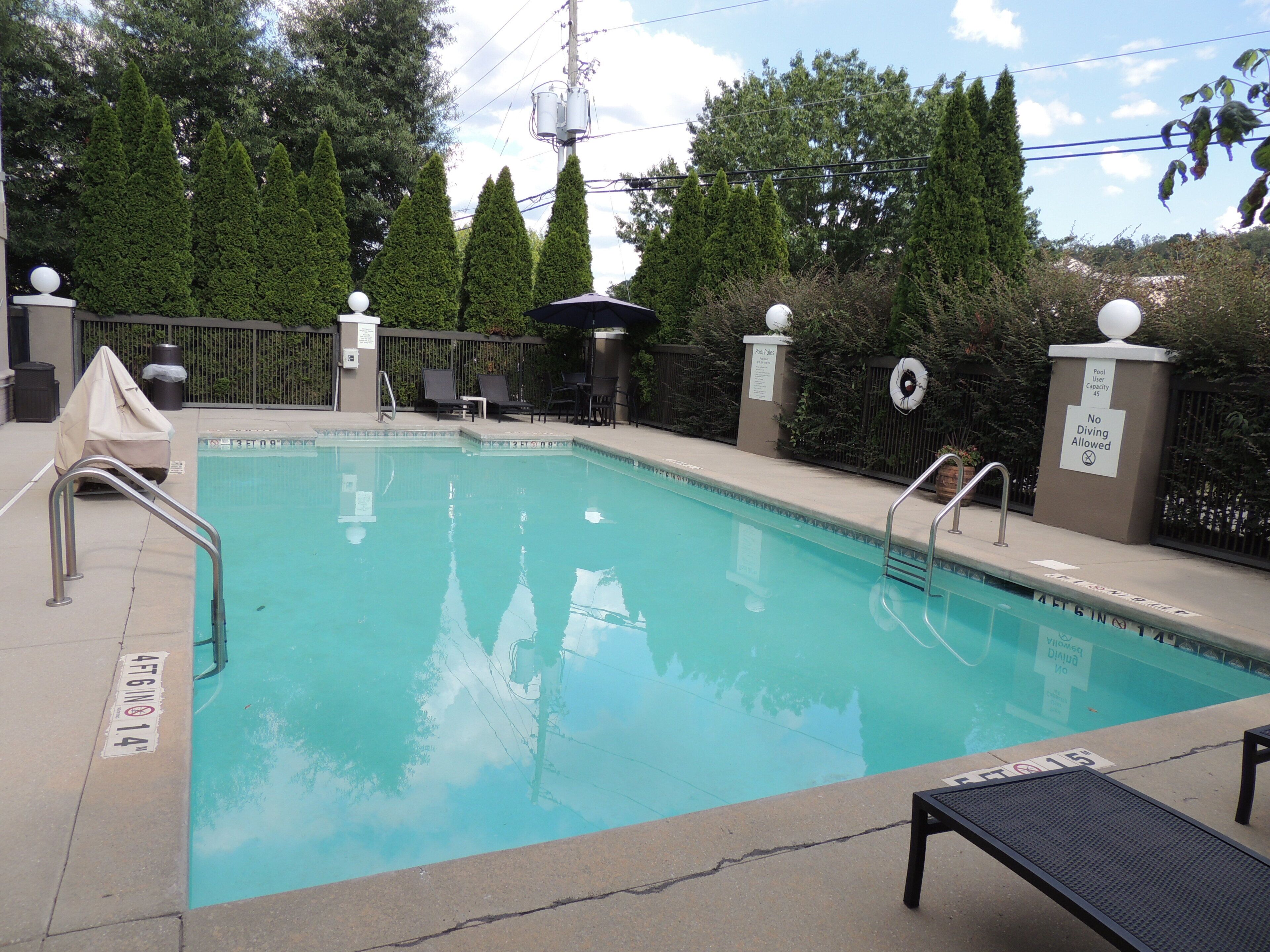 A rectangular pool with a clear blue water surface, surrounded by a low concrete deck. There are metal handrails on the pool's edge, and a lifebuoy attached to the wall. The pool area is enclosed by a metal fence with decorative posts topped with spherical objects.