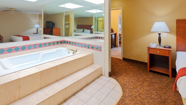 Un baño de hotel con una bañera grande, un espejo y una mesita de noche de madera con una lámpara.