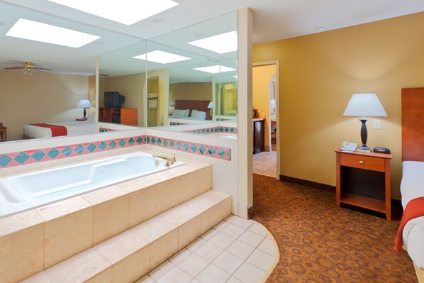 A hotel bathroom with a large bathtub, a mirror, and a wooden nightstand with a lamp.