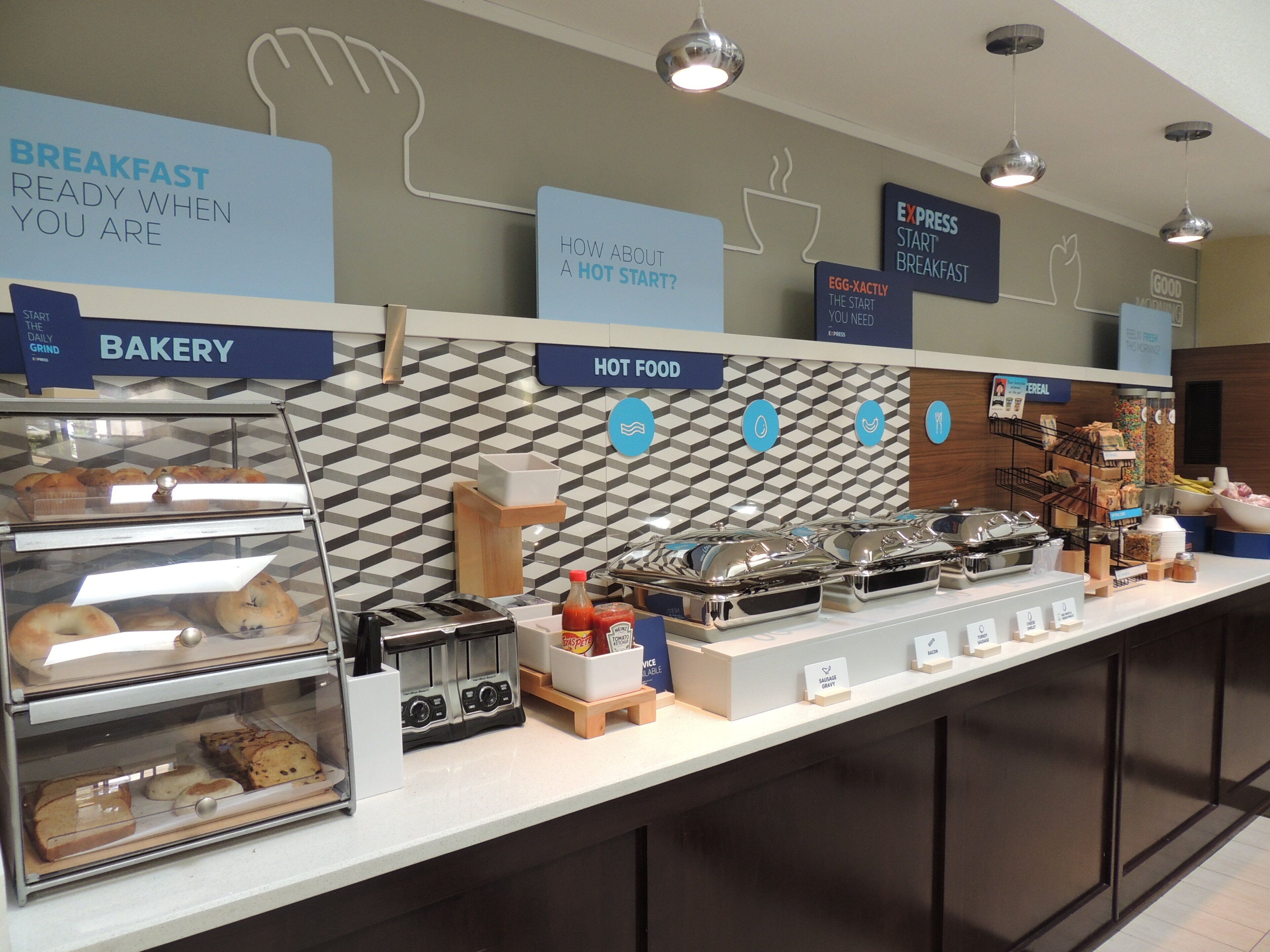 Zona de desayuno tipo buffet con secciones identificadas para panadería, comidas calientes y desayuno rápido.