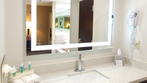 Baño de hotel con un espejo grande, un lavamanos, un secador de pelo y artículos de aseo en la encimera.