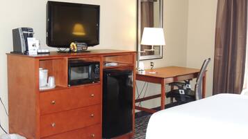 A hotel room with a wooden desk, a television, a microwave, a refrigerator, a lamp, and a bed.