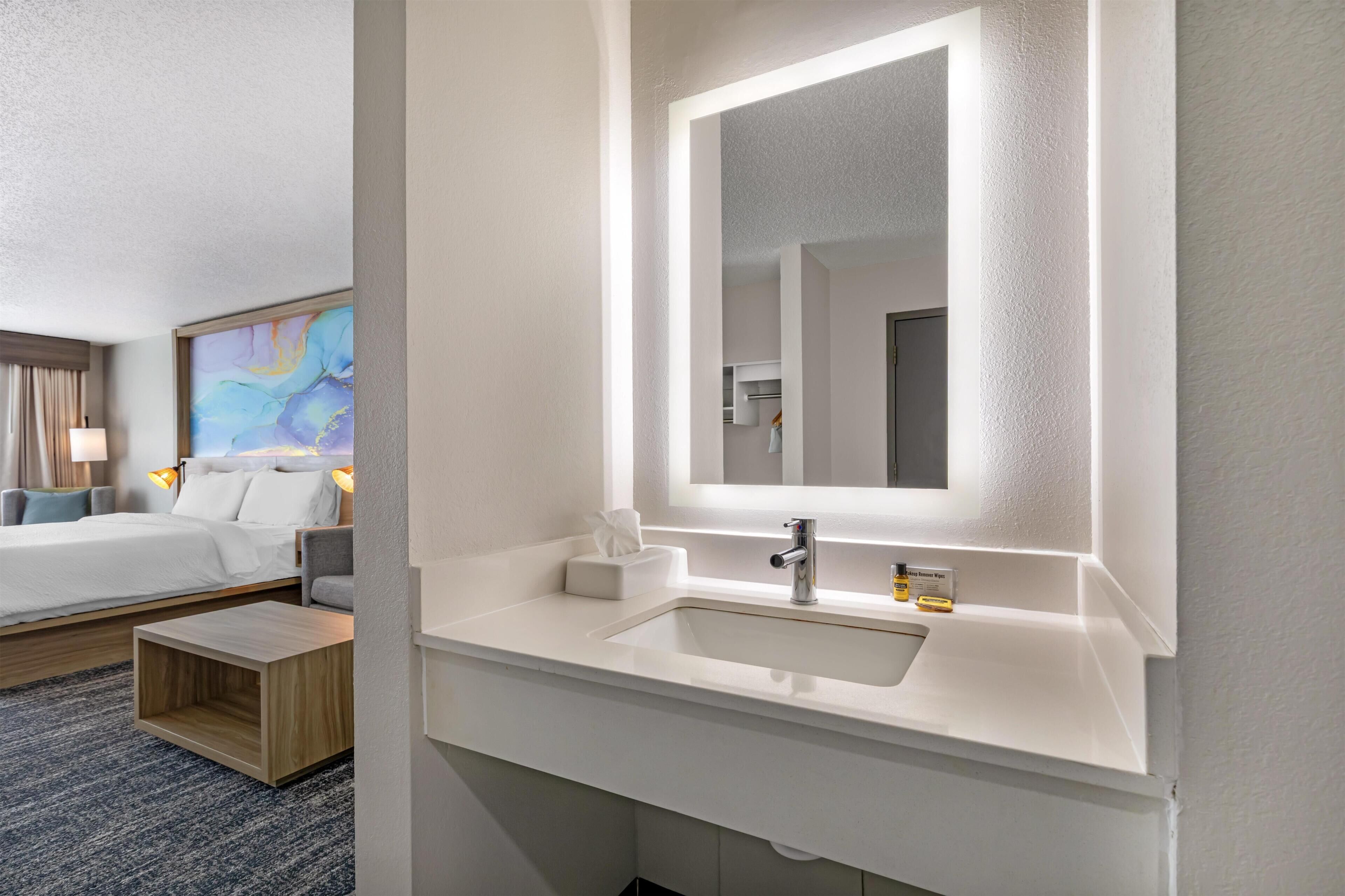 Une chambre d’hôtel moderne avec un lavabo blanc, un miroir, une table de chevet, un lit et un tableau au mur.