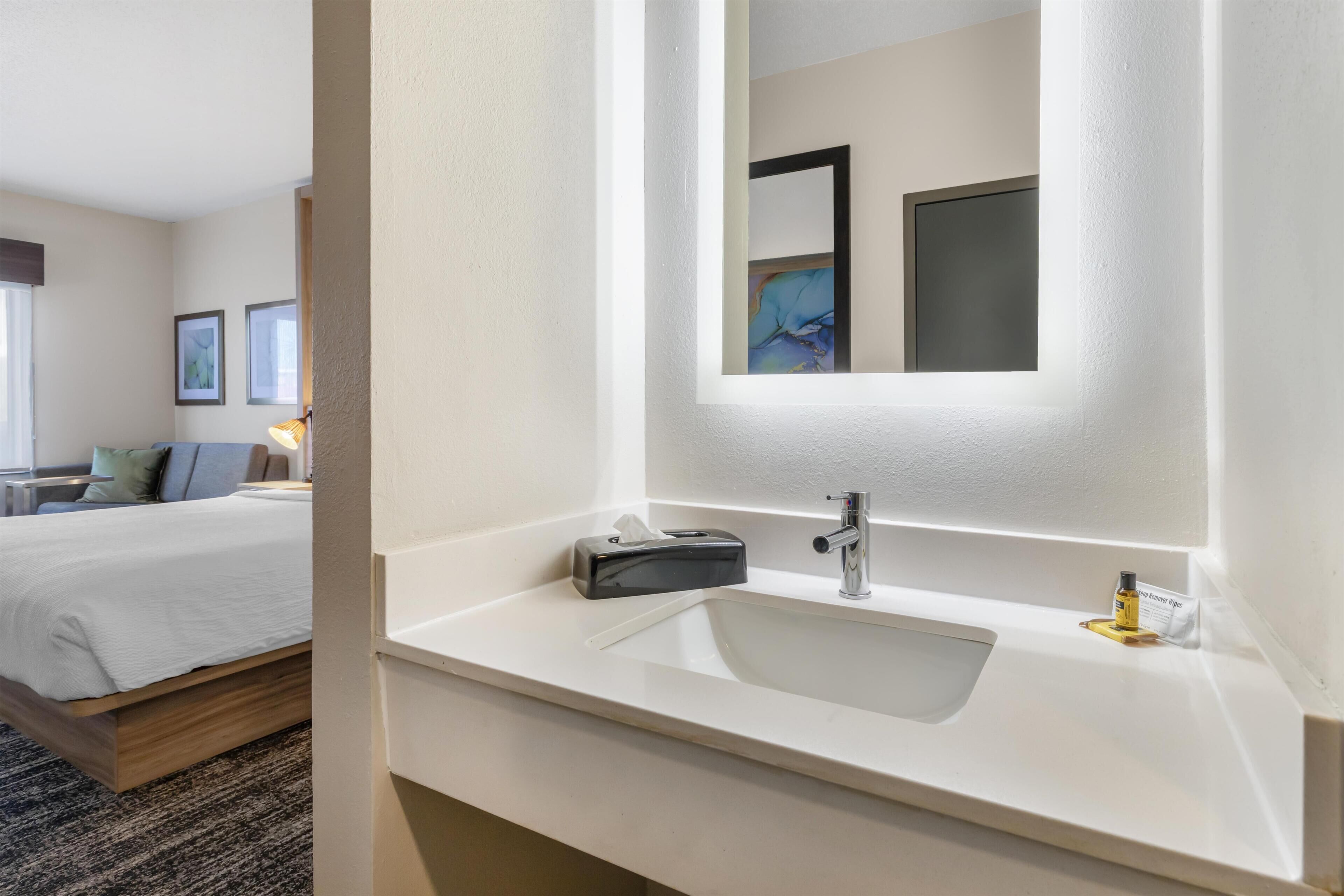 Un baño de hotel con un lavamanos blanco, un espejo y una mesita de noche con una lámpara.