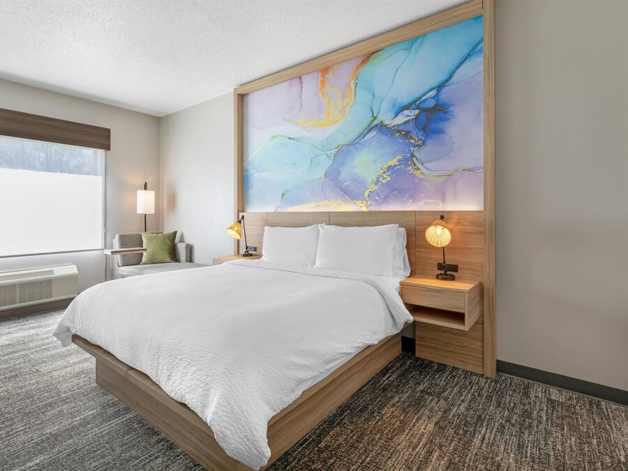 Ein Hotelzimmer mit einem großen Bett, Nachttischlampen, einem Nachttisch, einem Fenster mit Jalousien und einem an der Wand hängenden abstrakten Gemälde.