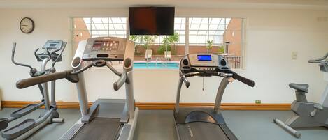 Un gimnasio con cintas de correr, elíptica y bicicleta estática. Hay un ventanal con vistas a una piscina y un reloj en la pared.