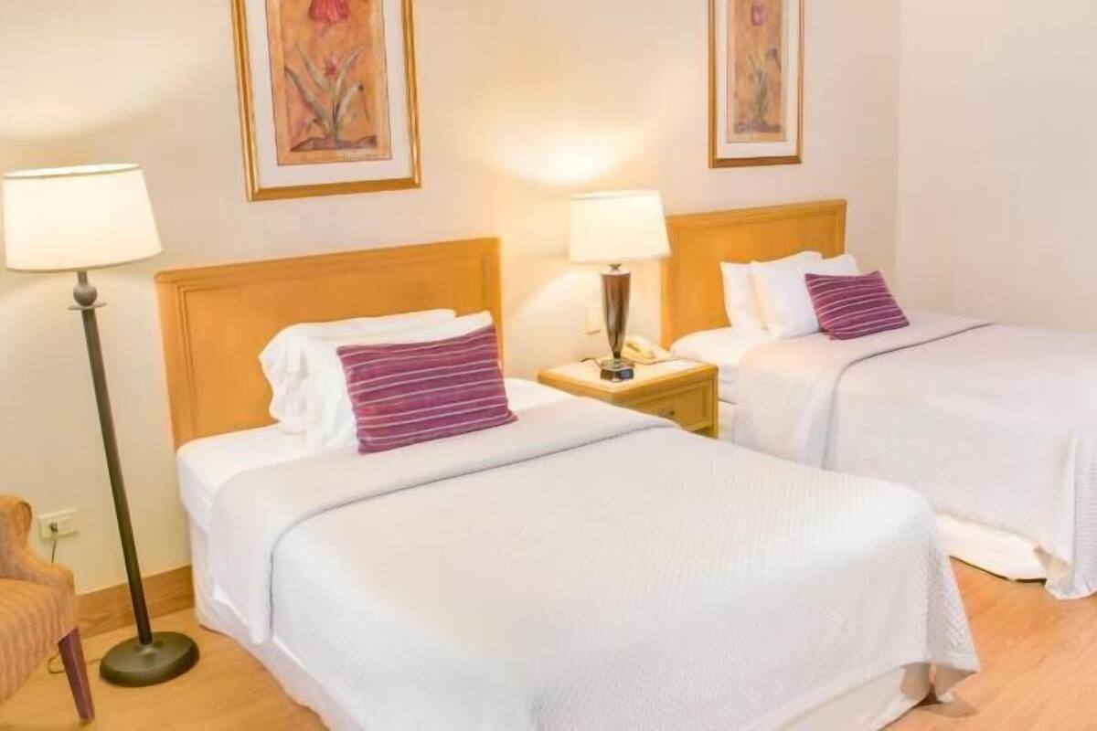 Habitación de hotel con dos camas, una mesita de noche con lámpara, una silla y obras de arte en las paredes.