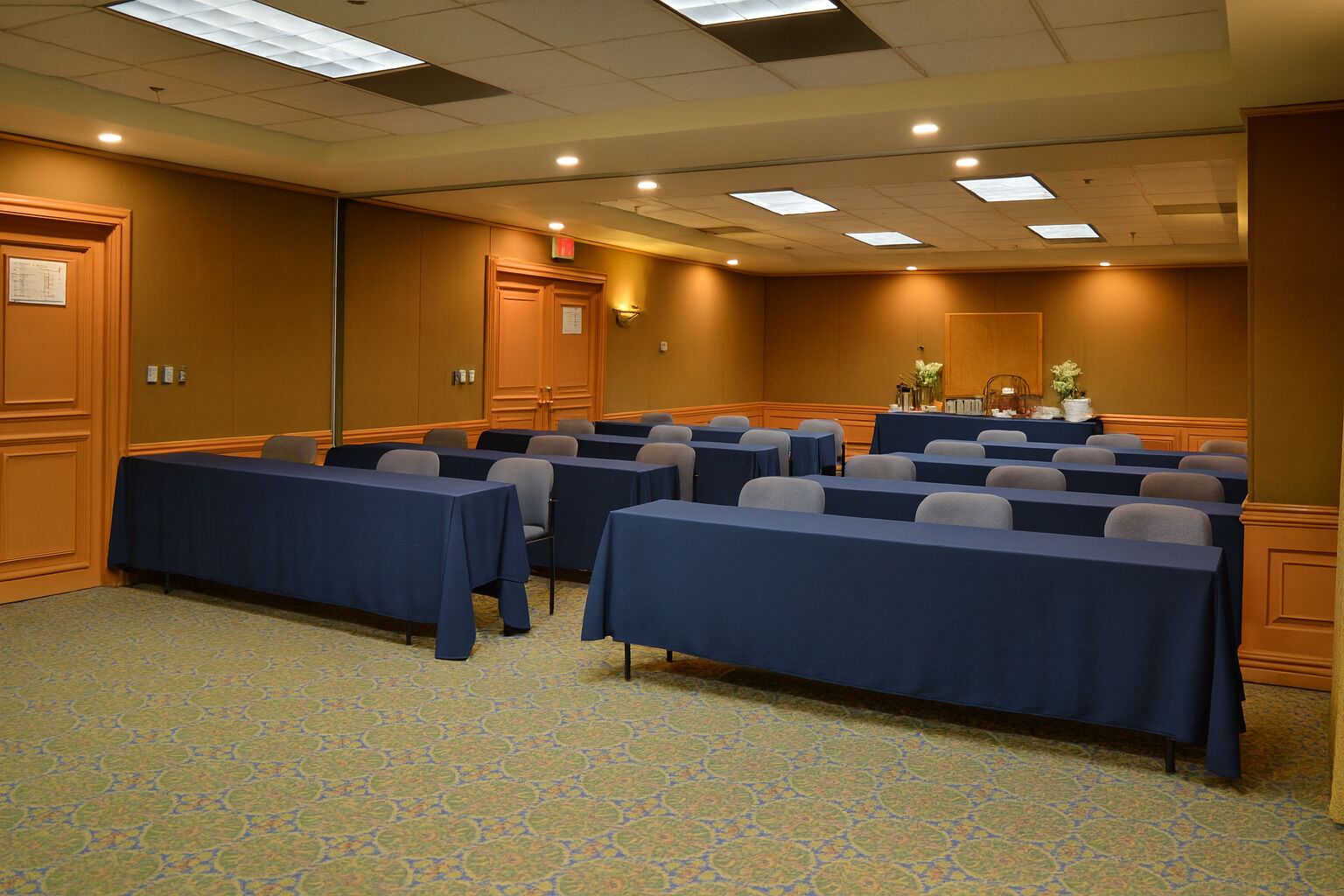 A conference room with rows of tables and chairs, equipped with overhead lighting and a patterned carpet.