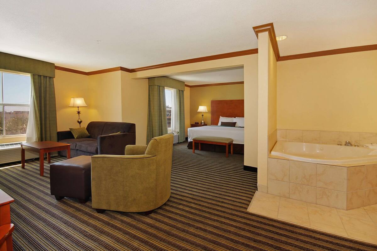 Een hotelkamer met een groot bed, een zithoek met een bank en fauteuil, een kleine tafel, een bankje en een aparte badkamer met een bad.